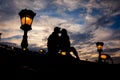 The silhouette of the sensual couple tenderly rubbing noses while sitting on the Chain Bridge near lightning street lamp
