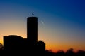 Silhouette of skyscraper with flag on top against a gradient twilight sky with turbojet trails