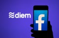 Silhouette of smartphone with Facebook on the screen and Facebook Diem crypto coin logo on the large blurred screen.