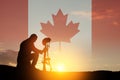 Silhouette of soldier kneeling with his head bowed on a background of sunset or sunrise and Canada flag.