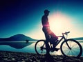 Silhouette of sportsman holding bicycle on lake beach, colorful sunset cloudy sky in background Royalty Free Stock Photo