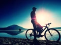 Silhouette of sportsman holding bicycle on lake beach, colorful sunset cloudy sky in background Royalty Free Stock Photo