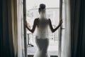 Silhouette of stylish bride opening window balcony in soft light in hotel room. Back of gorgeous sensual bride in white gown.