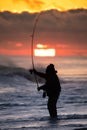 Silhouette of a surf caster fishing on the beach during a vibrant sunset.