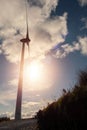 Silhouette of a tall wind generator against blue cloudy sky. Renewable source of energy in a plain field. Sun flare