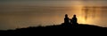 Silhouette of two girls and a puppy sitting on the beach against a background of bright orange water space lit by the Royalty Free Stock Photo