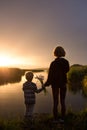 silhouette of woman and child holding each other\'s hands stand with backs near lake against backdrop of fiery sunset Royalty Free Stock Photo