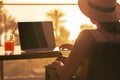 Silhouette of woman freelancer in straw hat drinking juice during working on laptop sitting on the armchair near table on balcony Royalty Free Stock Photo