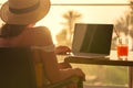 Silhouette of woman freelancer in straw hat drinking juice during working on laptop sitting on the armchair near table on balcony Royalty Free Stock Photo