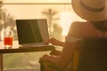Silhouette of woman freelancer in straw hat drinking juice during working on laptop sitting on the armchair near table on balcony. Royalty Free Stock Photo