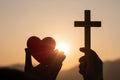 Silhouette of woman hands praying with cross and holding a red heart ball in nature sunrise background, Crucifix, Symbol of