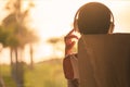 Silhouette of woman with headphones sitting on deck chair on terrace and looking on palm trees sea beach at sunset.