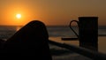 Silhouette of a woman`s foot drinking coffee in the open air at the sunrise, on the seashore