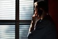 Silhouette of young attractive man talking on mobile phone while standing in dark room at shadow blinds