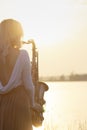 Silhouette of a young beautiful girl playing the saxophone at sunrise by the river, a woman in a long dress on the nature at