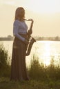 Silhouette of a young beautiful girl playing the saxophone at sunrise by the river, a woman in a long dress on the nature at