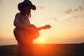 Silhouette of young free woman in straw hat playing country music on a guitar at sunset