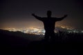 Silhouette of a young man with raised-up arms, standing on the top of the mountain under night sky with stars and near city. Royalty Free Stock Photo