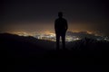 Silhouette of a young man standing on the top of the mountain under night sky with stars and near city. Royalty Free Stock Photo