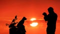 Silhouette young man using camera to taking picture of his pets [puppy and 2 parrots] on motorcycle with blurred colorful sunset