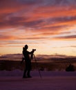 Silhouette of a young photographer shooting a snowy landscape at sunset with a camera on a tripod Royalty Free Stock Photo