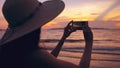 Silhouette of young tourist woman in hat taking photo with cellphone during sunset in ocean beach Royalty Free Stock Photo