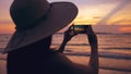 Silhouette of young tourist woman in hat taking photo with cellphone during sunset in ocean beach Royalty Free Stock Photo