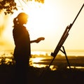 Silhouette of a young woman painting a picture with paints on canvas on an easel outdoors, girl profile engaged in art at sunset