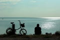 Silhouettes of bicycle and man sitting at the seashore and looking at sea
