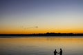 Silhouettes of man and woman fishing with sunset reflecting in water and flock of birds flying in formation overhead Royalty Free Stock Photo