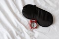 Silk black sleep mask, red clock, showing 6 o`clock on white rumpled sheets. Top view, flat lay. Horizontal. Concept of rest,