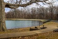 A silky green lake surrounded by a smooth concrete footpath, yellow winter grass and bare winter trees with gray sky