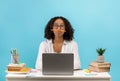 Silly young black female student sitting at desk with books and laptop, playing game with pencil on blue studio