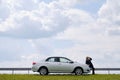 A Silver Car Is Parked On The Side Of A Lonely Road In A Rural Area, Against A Blue Sky With Clouds And A Green Field. Nearby, A