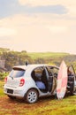 A silver grey car on a cliff with bright pink sirfboard and thermos on roof with its doors open. Surfing lifestyle on