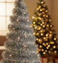Silver tinsel Christmas tree adorned with small white lights dominates the foreground. In the blurred background. Royalty Free Stock Photo