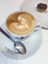 A simple cappuccino with beautiful coffee art and design, served in a white mug with side a silver spoon