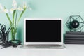 Simple office interior with blank laptop monitor, black stationery, books, abstract modern sculpture, white flowers in green mint. Royalty Free Stock Photo