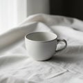 A simple white ceramic cup sits on a smoothly textured white fabric, possibly a tablecloth or bed Royalty Free Stock Photo