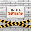 Simple white sign with text `Under Construction` hanging on a gray brick wall with warning tapes. Engineering concept