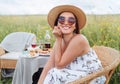 Sincerely smiling at camera young Woman in sunglasses and straw hat dressed light summer dress sitting in rattan chair on the