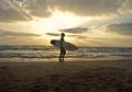 Single male surfer with a surfboard walking on a sandy beach on a cloudy sunset