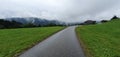 Single road between green fields with very cloudy, mysterious hanging clouds over the far away mountains