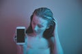 Single sad teen holding a mobile phone lamenting sitting on the bed in her bedroom with a dark light in the background Royalty Free Stock Photo