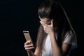 Single sad teen holding a mobile phone lamenting sitting on the bed in her bedroom with a dark light in the background