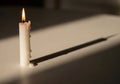 A single white candle stands upright on a flat surface, casting a long shadow. The candle is lit, Royalty Free Stock Photo