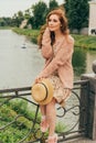 sits on a fence, in the hands of a hat, a beige dress in a flower and a beige sweater.beautiful red-haired girl.picture against th Royalty Free Stock Photo