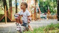 Sits on the green grass. Cheerful little girl on roller skates have a good time in the park near attractions Royalty Free Stock Photo