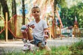 Sits on the green grass. Cheerful little girl on roller skates have a good time in the park near attractions Royalty Free Stock Photo