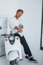 Sits on motorcycle. Caucasian young guy in elegant white shirt indoors at home